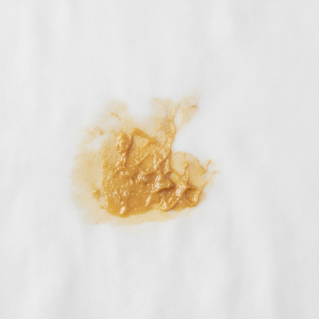 A detailed, professional studio photo capturing a close-up of a pristine white cotton t-shirt severely marred by a prominent, yellowish, oily sunscreen stain. The stain appears deeply embedded within the fabric weave, highlighting its stubborn nature and the challenge it presents. The surrounding fabric is crisp and clean, creating a strong visual contrast with the discolored area. The lighting is bright and even, accentuating the texture of the fabric and the greasy sheen of the stain, making it look tangible and visually impactful. The background is softly blurred to keep focus on the stained garment.
