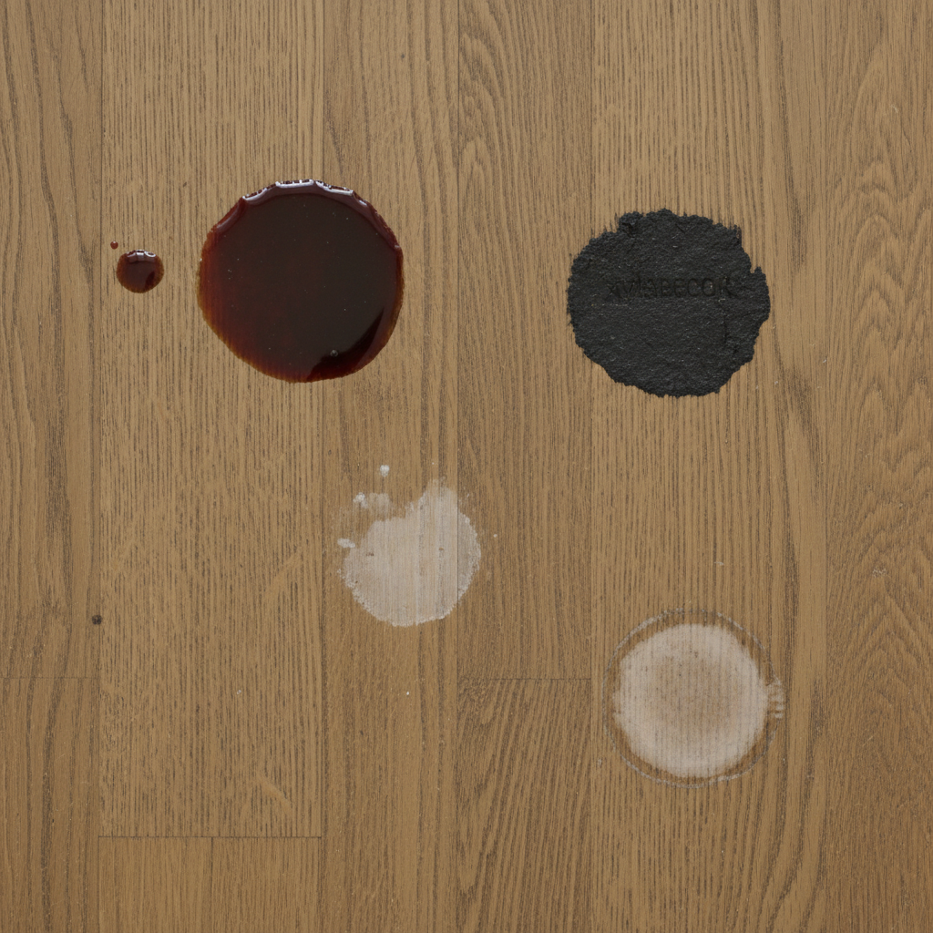 A top-down, slightly zoomed-in professional photograph of a wooden surface, like a part of a floorboard or tabletop, clearly displaying different types of Xyladecor-related stains. The surface should feature a fresh, wet-looking splatter of wood stain, a distinct, darker patch of dried-on stain, a subtle, faded discolored area representing an old stain, and a visible water ring. The natural wood grain should be partially visible beneath the imperfections, highlighting the problematic nature of these blemishes.