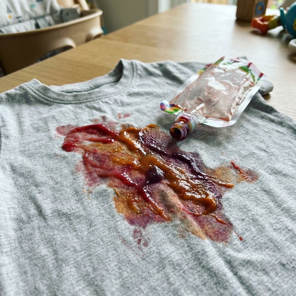 A close-up, slightly elevated shot of a vibrant, multi-colored (e.g., red, orange, purple hues) fruit puree stain on a light grey or white child's t-shirt. The stain appears wet, slightly sticky, and has spread a bit on the fabric. A small, empty, crumpled fruit puree pouch is casually tossed near the stain, indicating the source. The background is softly blurred, hinting at a domestic, slightly messy environment. Professional, clean product photography with natural lighting.